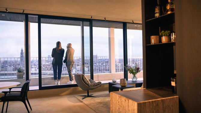 Alt: Innenansicht einer modernen Wohnung mit breiten Schiebetüren und zwei Personen auf einem Balkon.
