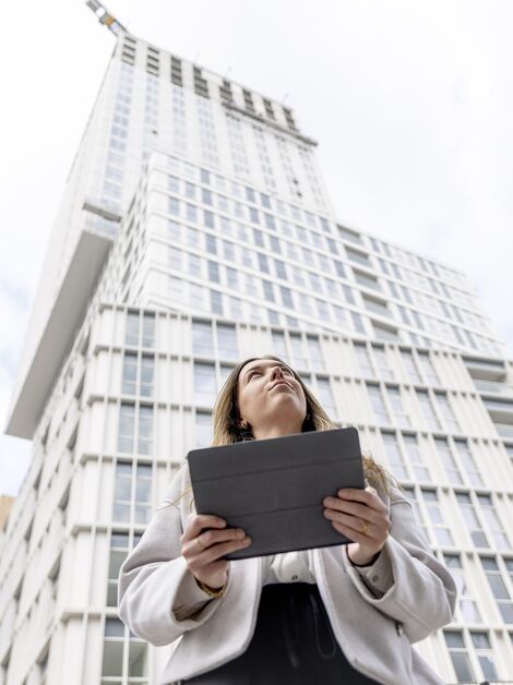 Blonde Frau mit iPad schaut nach oben in Richtung weißes Wohnhaus.